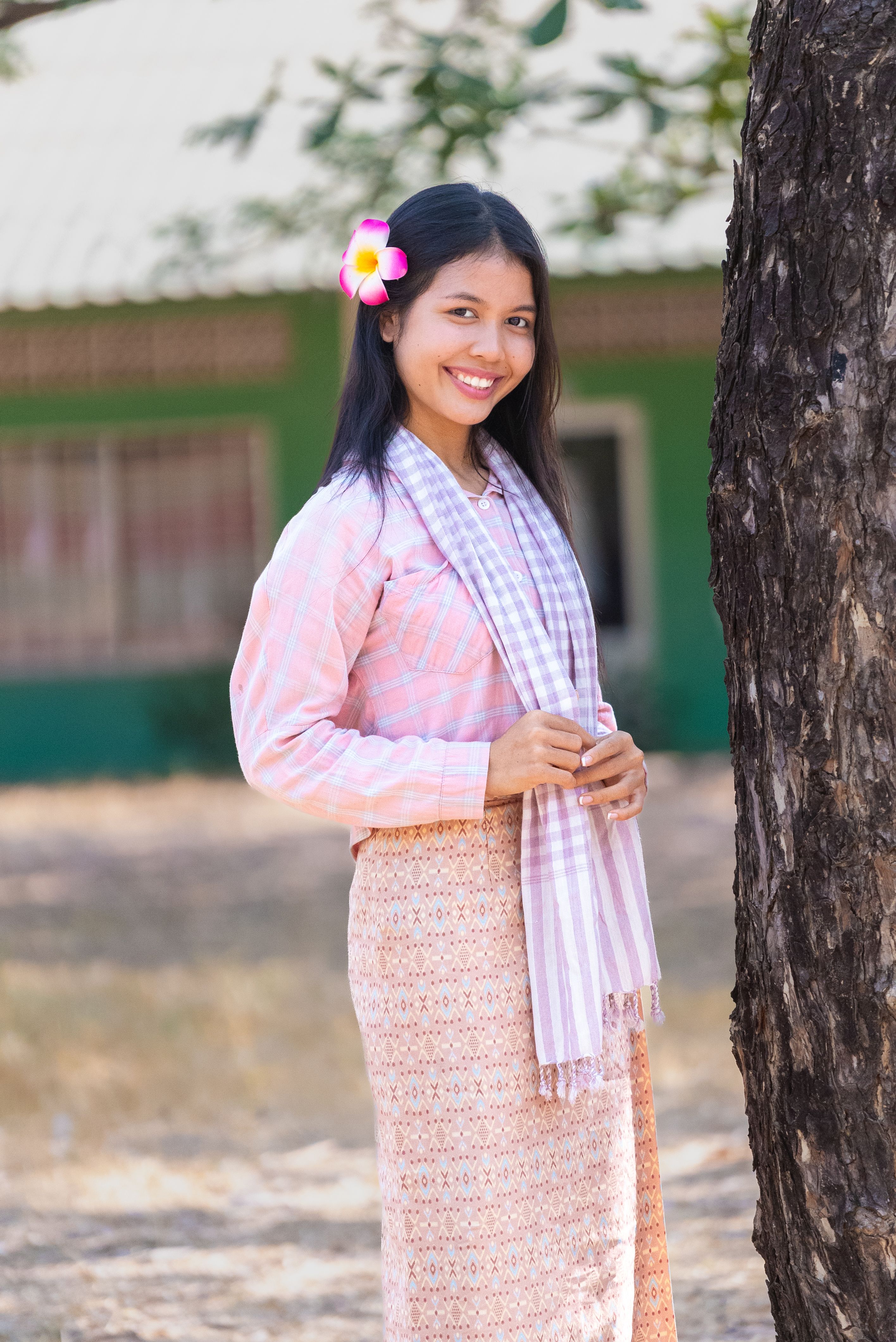 Happy woman wearing traditional attire with a flower in her hair, standing next to a tree.