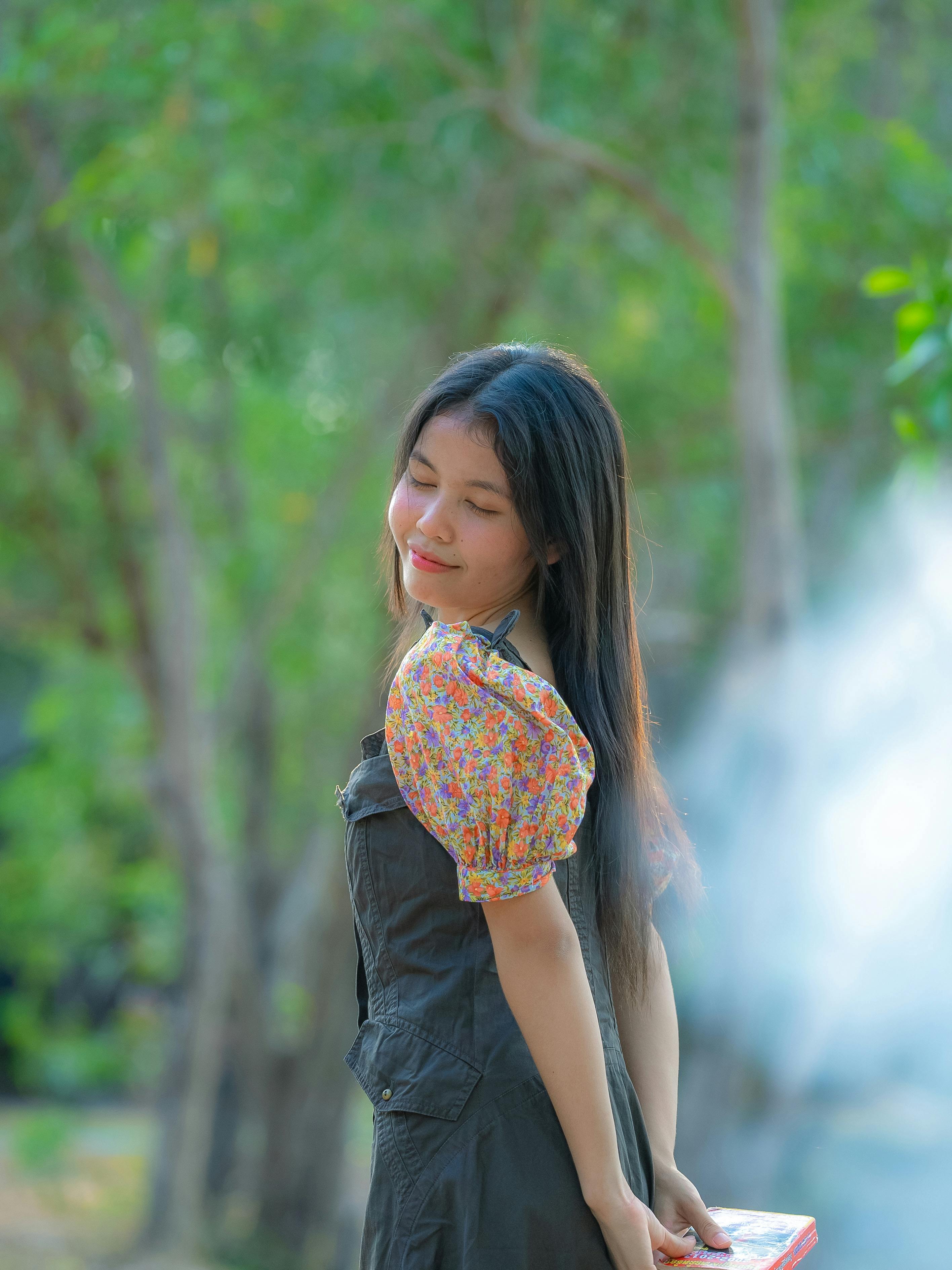 Безкоштовне Молода жінка в квітковій сукні ніжно посміхається, оточена туманним зеленим лісовим пейзажем. стокове фото