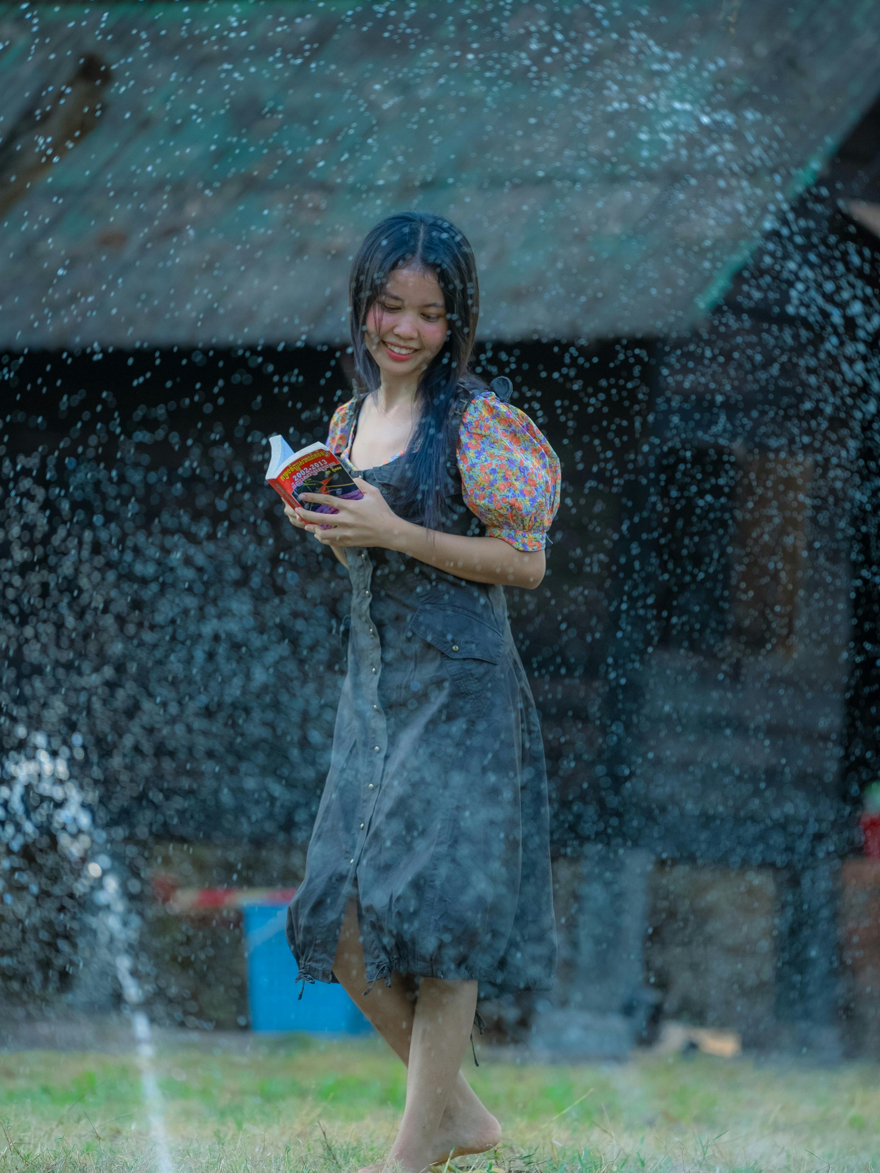 ingyenes Egy mosolygós nő élvezi az olvasást, miközben játékosan áll egy locsoló alatt egy rusztikus kültéri környezetben. Stockfotó