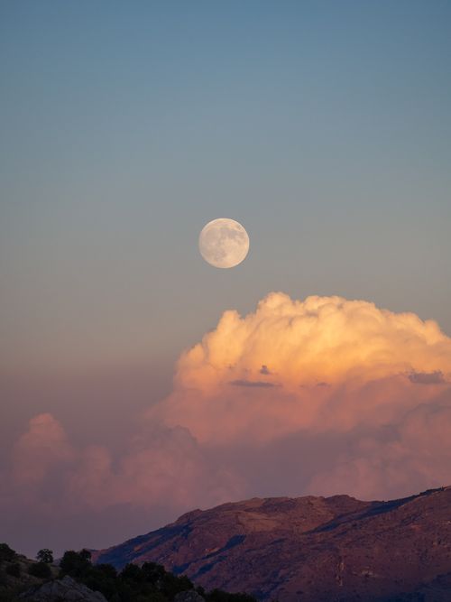 Free Beautiful full moon rising above pink clouds and mountain ridge at sunset, creating a serene and majestic view. Stock Photo