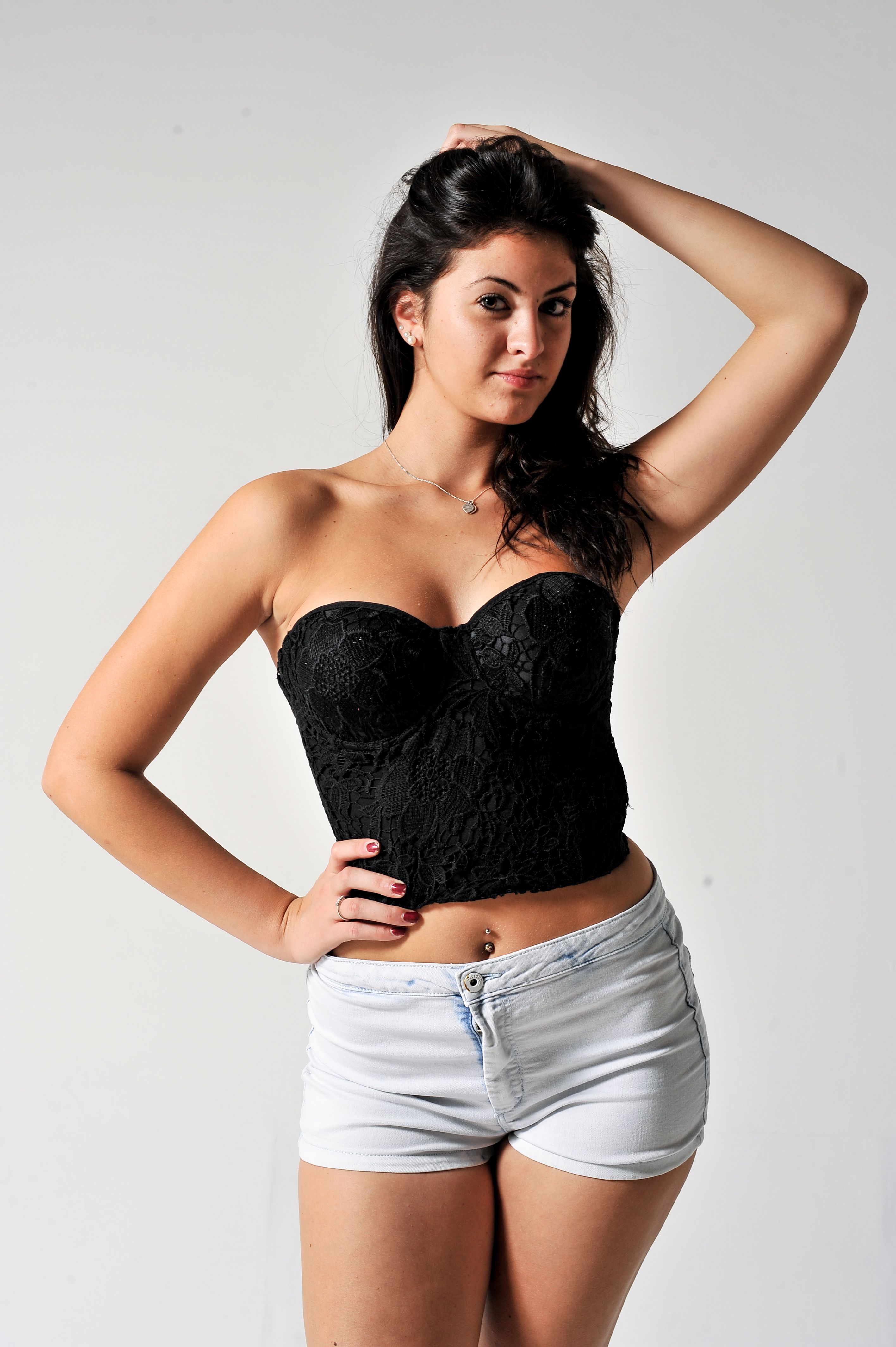 Confident young woman posing in black lace top and denim shorts against a plain background.