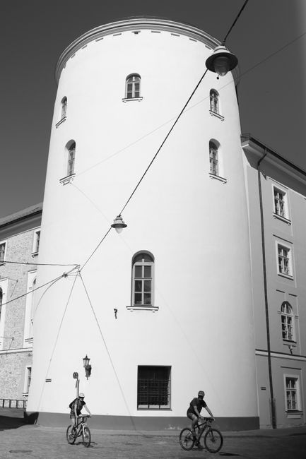 A black and white photo of cyclists passing an architectural building in Riga, Latvia.