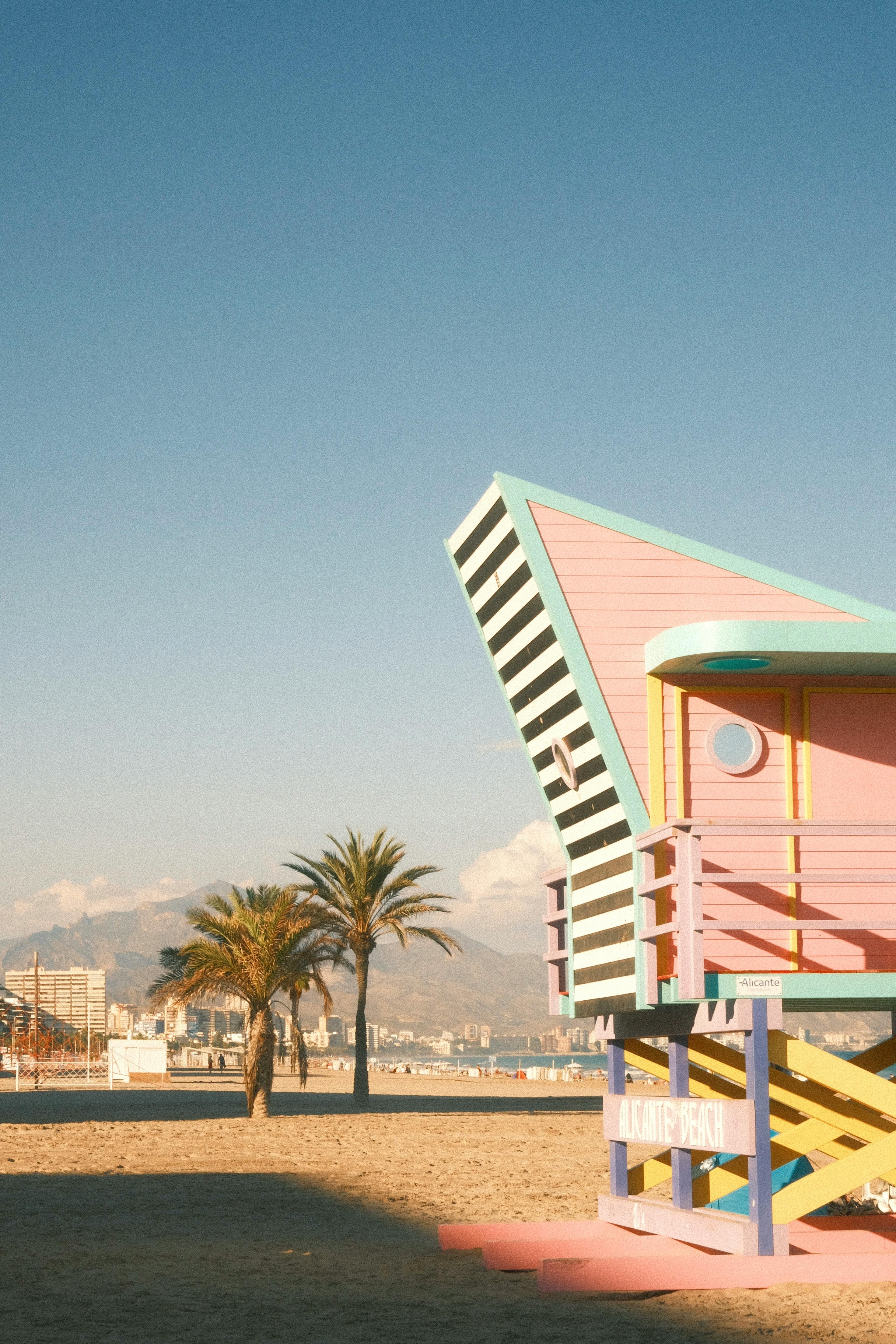 Vibrant lifeguard hut on a sandy beach with palm trees and mountains in the background.