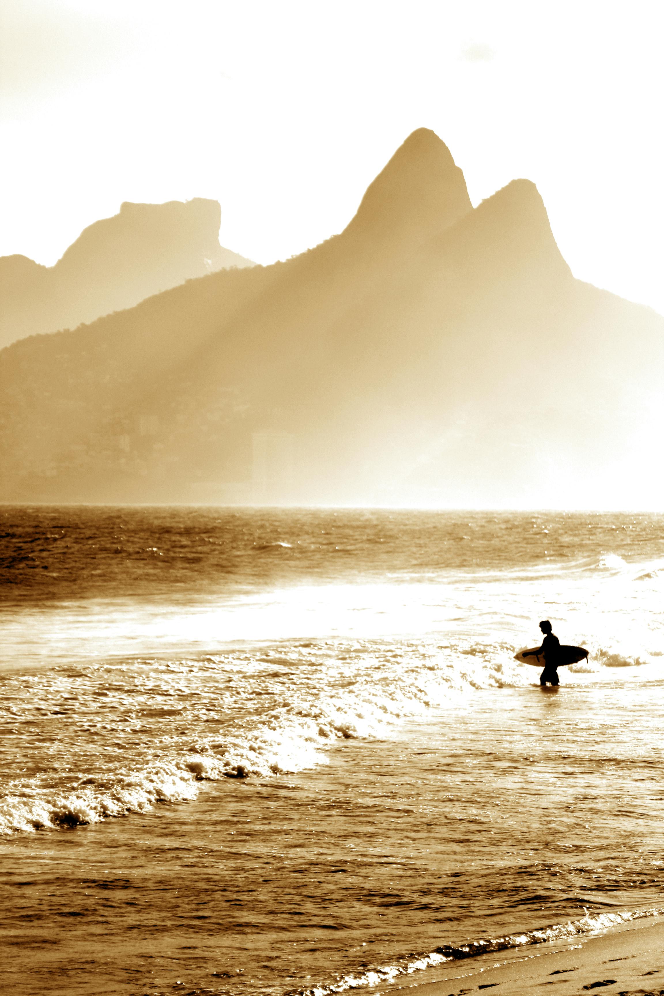 A lone surfer walks into the sunset with the iconic Dois Irmãos peaks in the background.