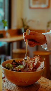 A hand holds a fork in a rustic bowl filled with salad and crispy bread in a cozy interior setting.