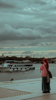 Woman standing by the Bosphorus river, watching a ferry boat with a picturesque Istanbul skyline.