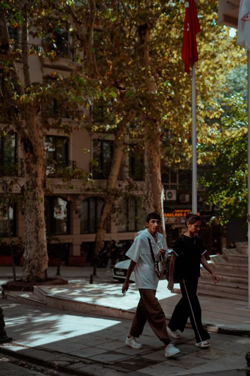 Free Two young adults stroll on a tree-lined street in a sunlit urban area, offering a cinematic feel. Stock Photo