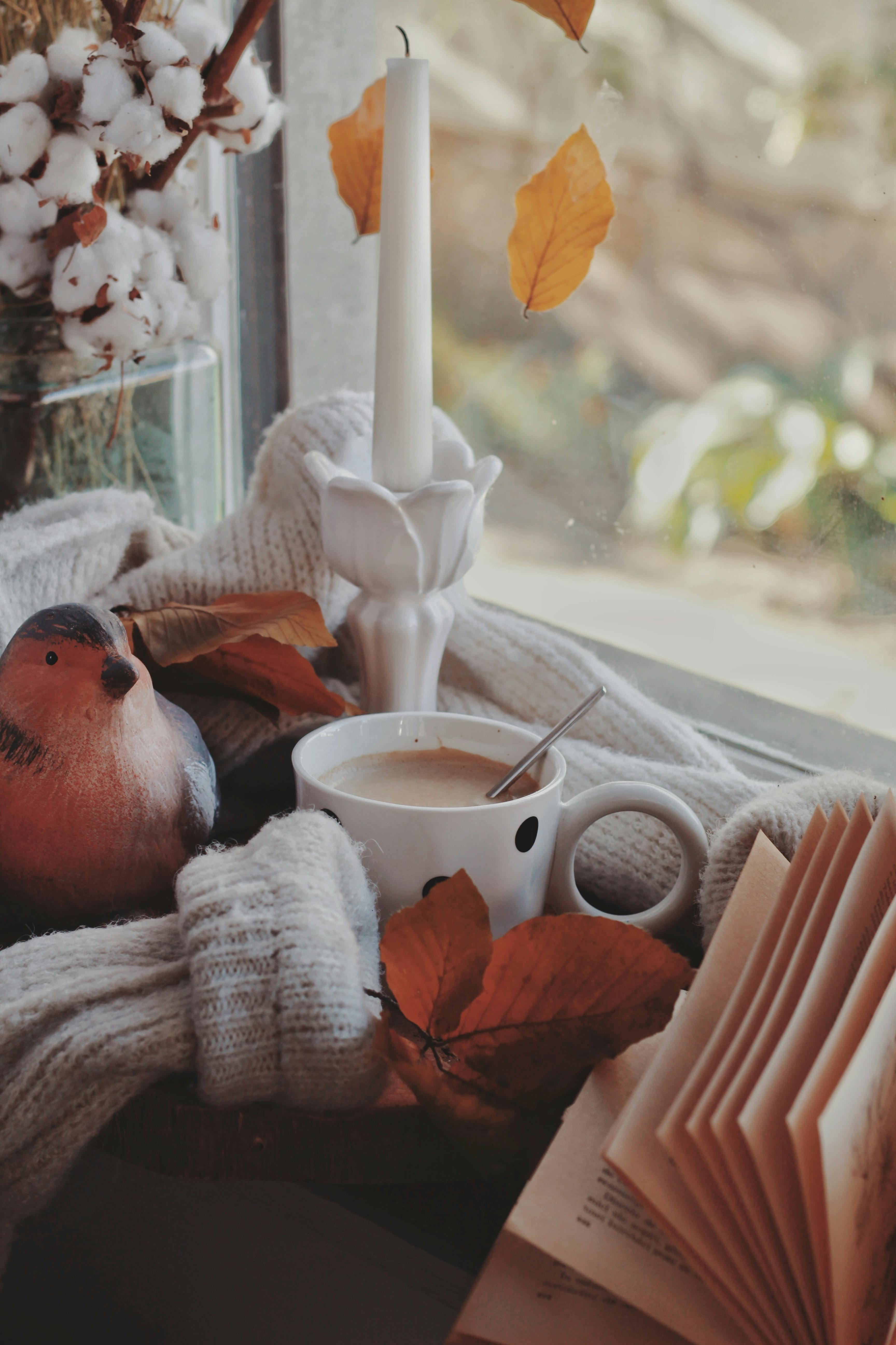 Warm autumn scene with coffee, candle, sweater, and leaves by the window.