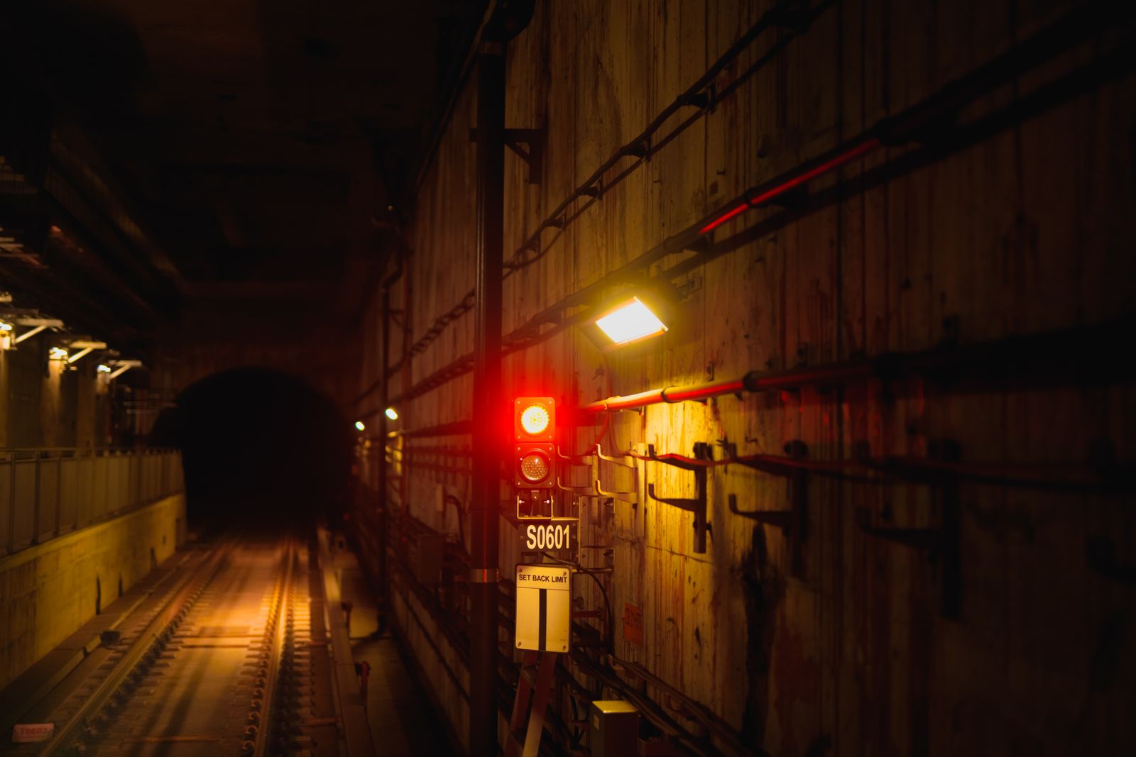 A dark subway tunnel with a lit red signal light on the right hand wall beside a yellow floodlight and a plate marked S0601, the rails on the left reflecting orange light into blackness ahead