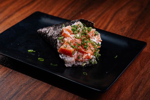 A close-up of a salmon sushi hand roll served on a black plate, garnished with green onions.