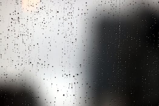 Raindrops trickle down a window with a blurred city skyline background, creating a moody atmosphere.