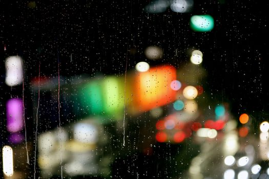 Raindrops on window create a bokeh effect with colorful city lights in the background.