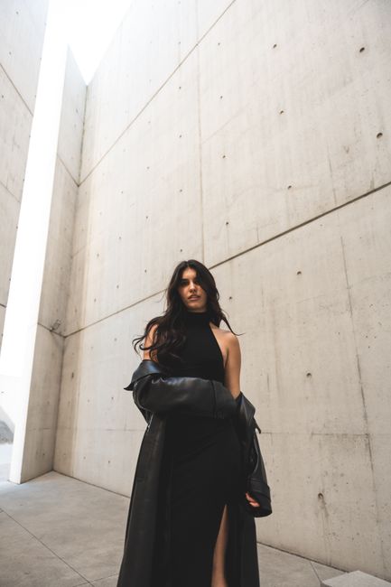 Fashionable woman in black dress poses dramatically against a modern concrete background.