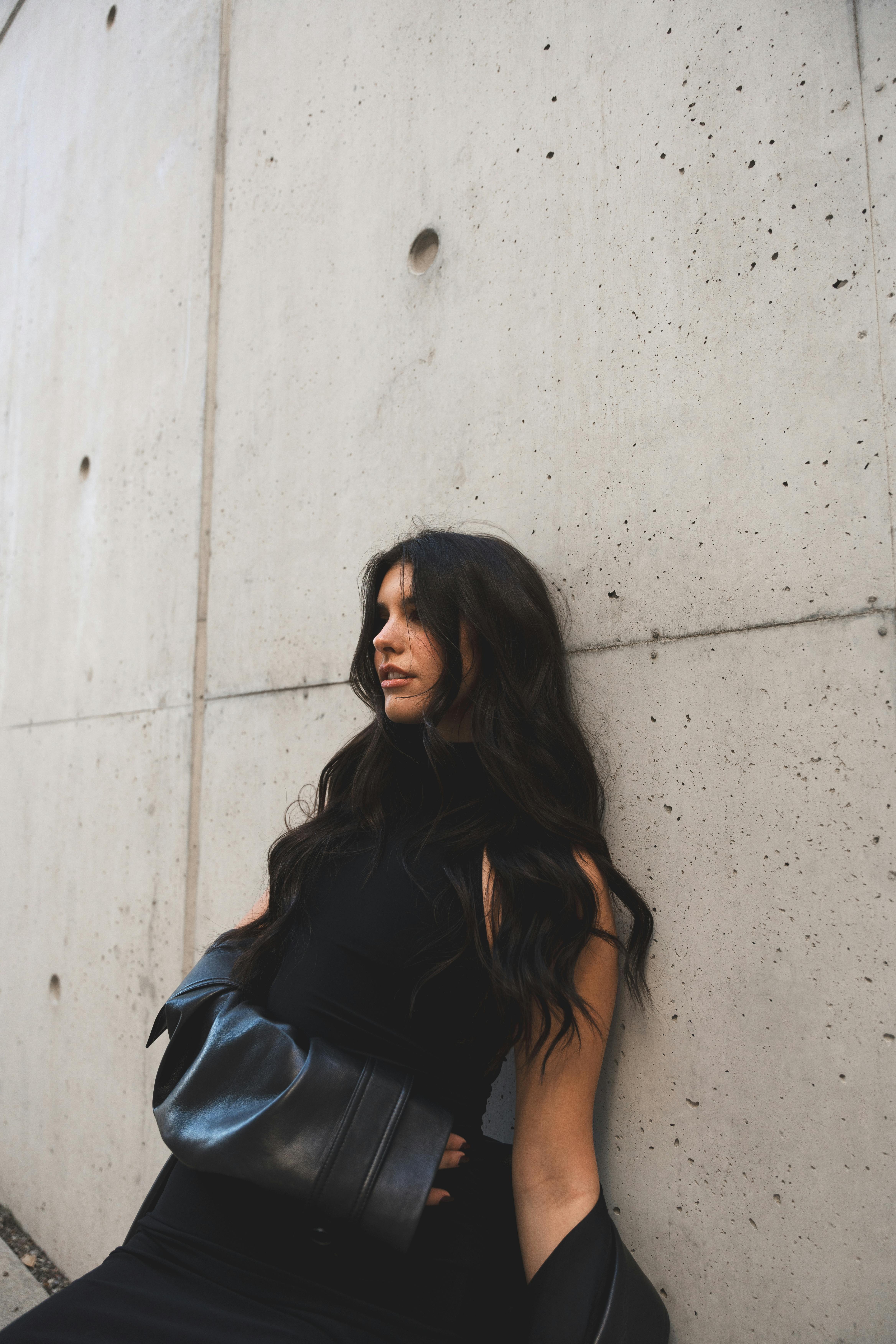 A fashionable woman in a black dress poses against a concrete wall, showcasing modern style.