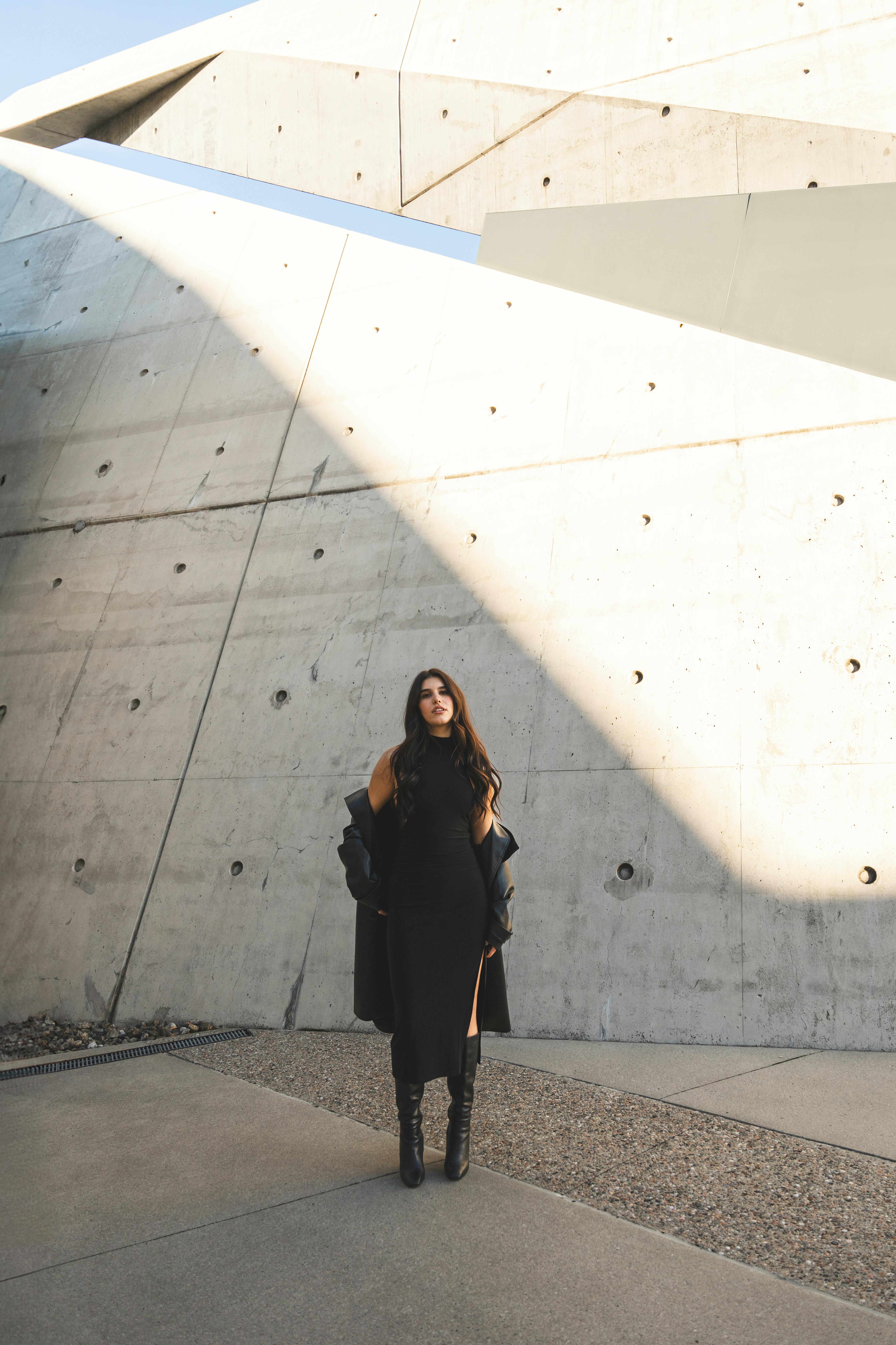 Woman in black dress poses stylishly against striking concrete architecture.