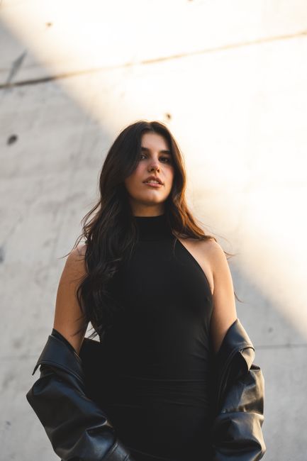Fashionable woman poses confidently in black dress with sunlight highlighting features against a modern concrete backdrop.