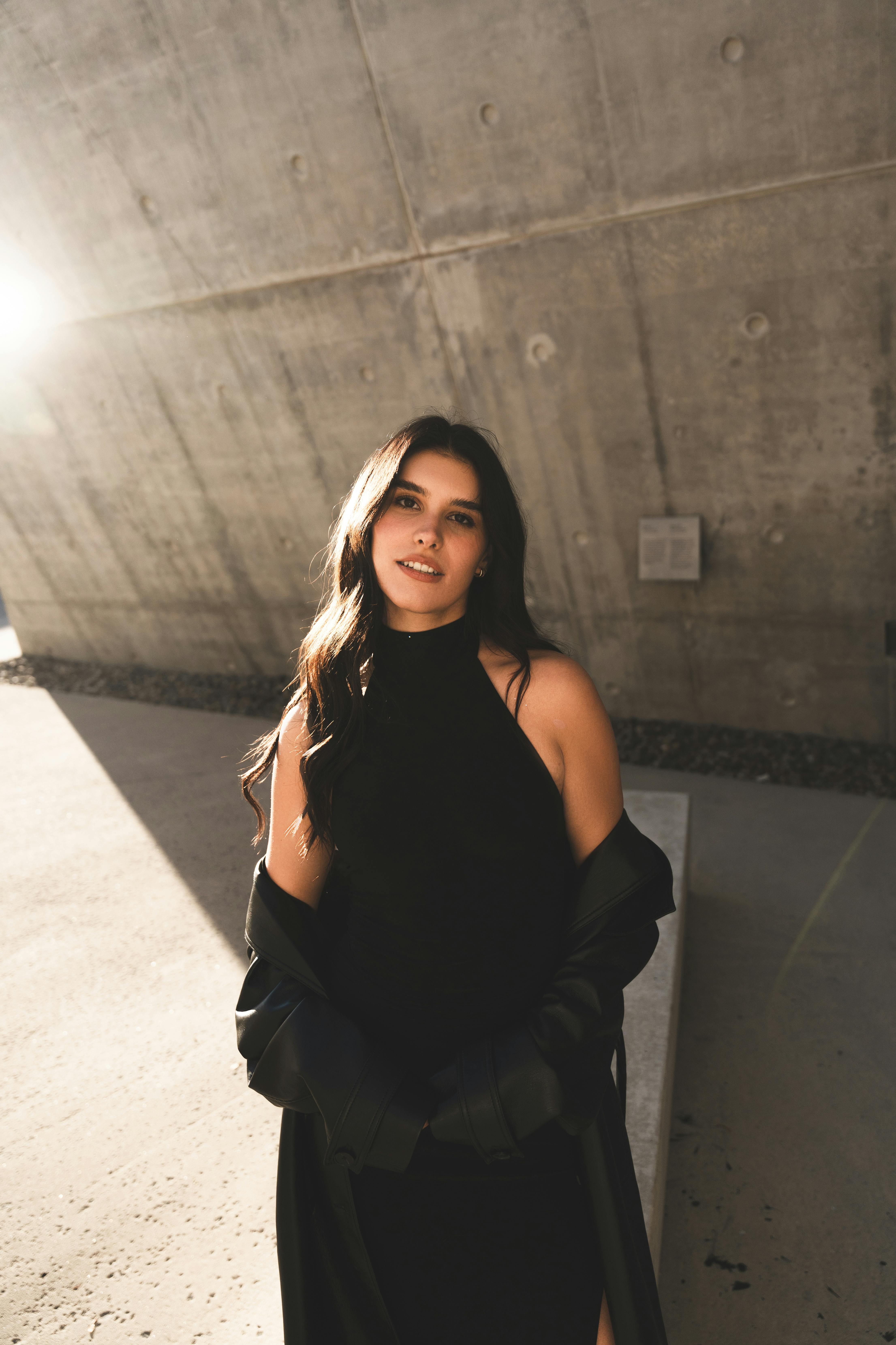 Elegant woman posing in black dress with sunlight in urban environment.