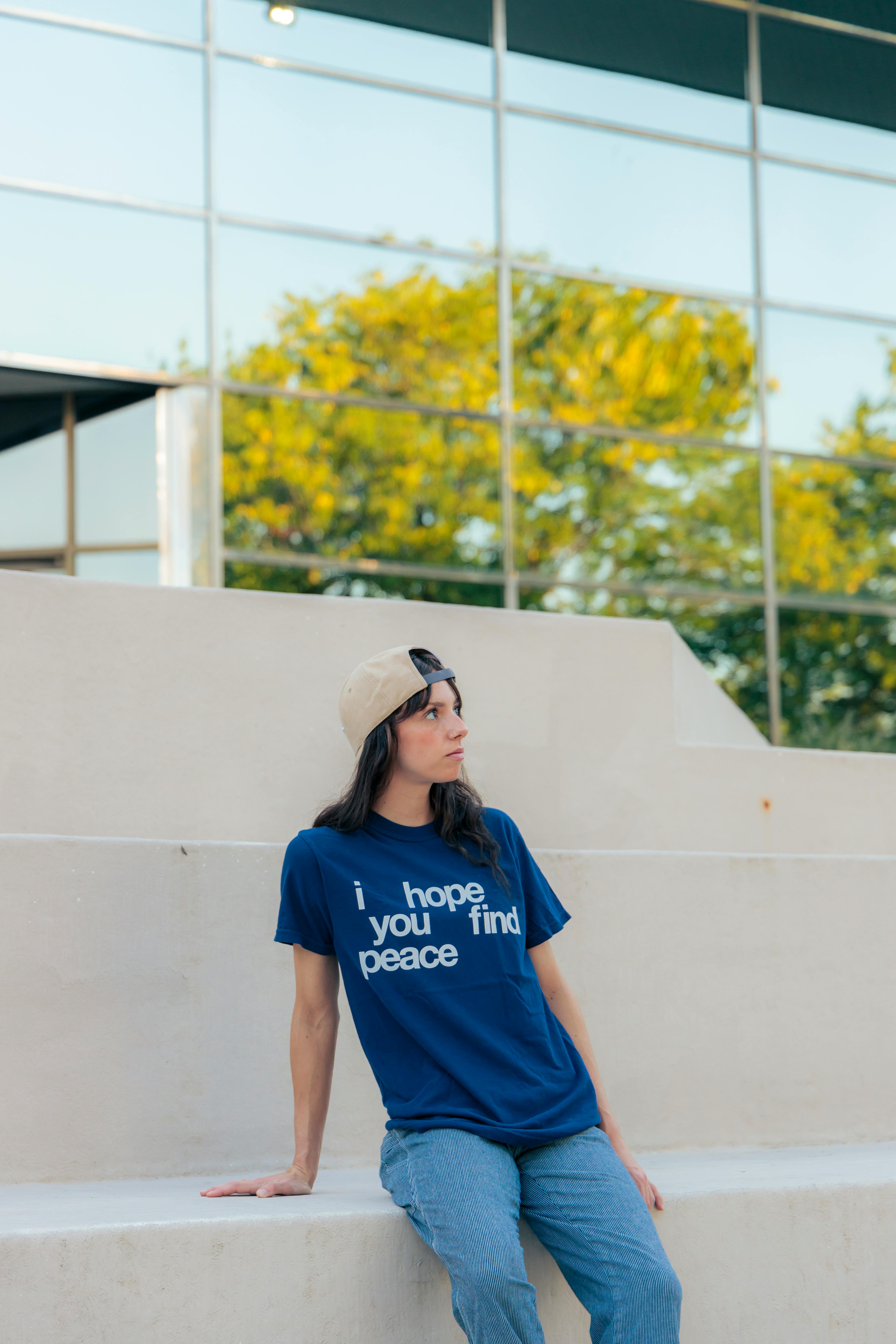 Woman in blue shirt with 'I hope you find peace' text, seated outdoors.