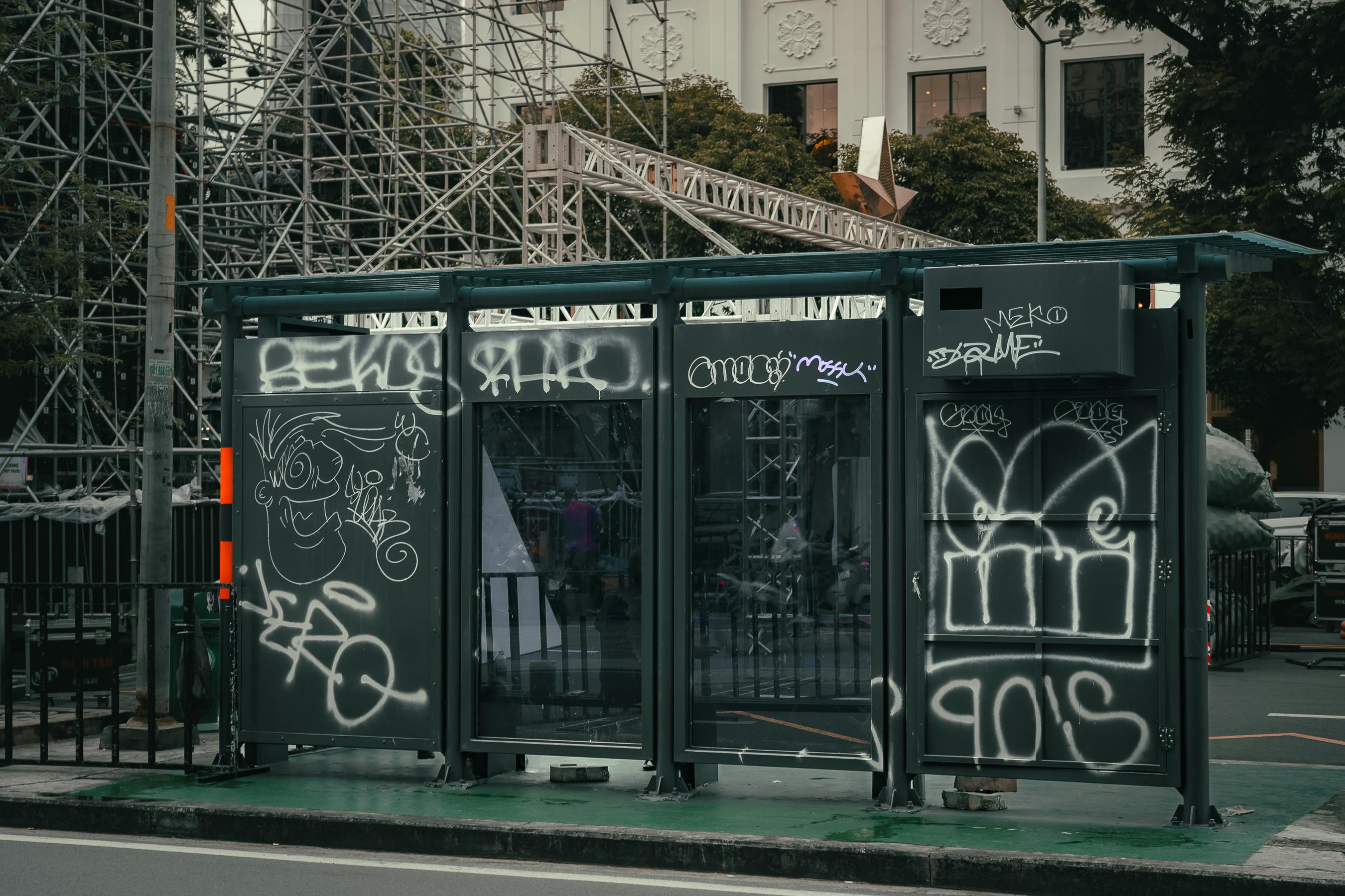 Bus stop with graffiti in an urban setting, surrounded by scaffolding and trees.