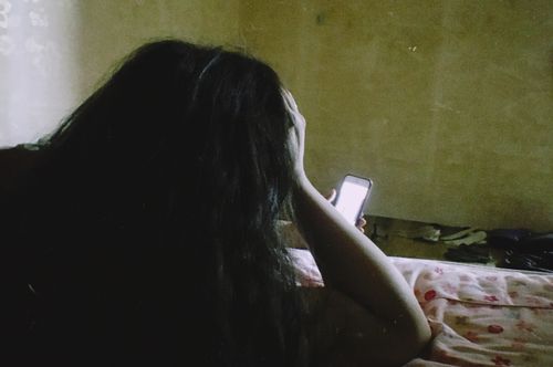 Free A young woman lies on a bed using her smartphone in a dimly lit room. Stock Photo