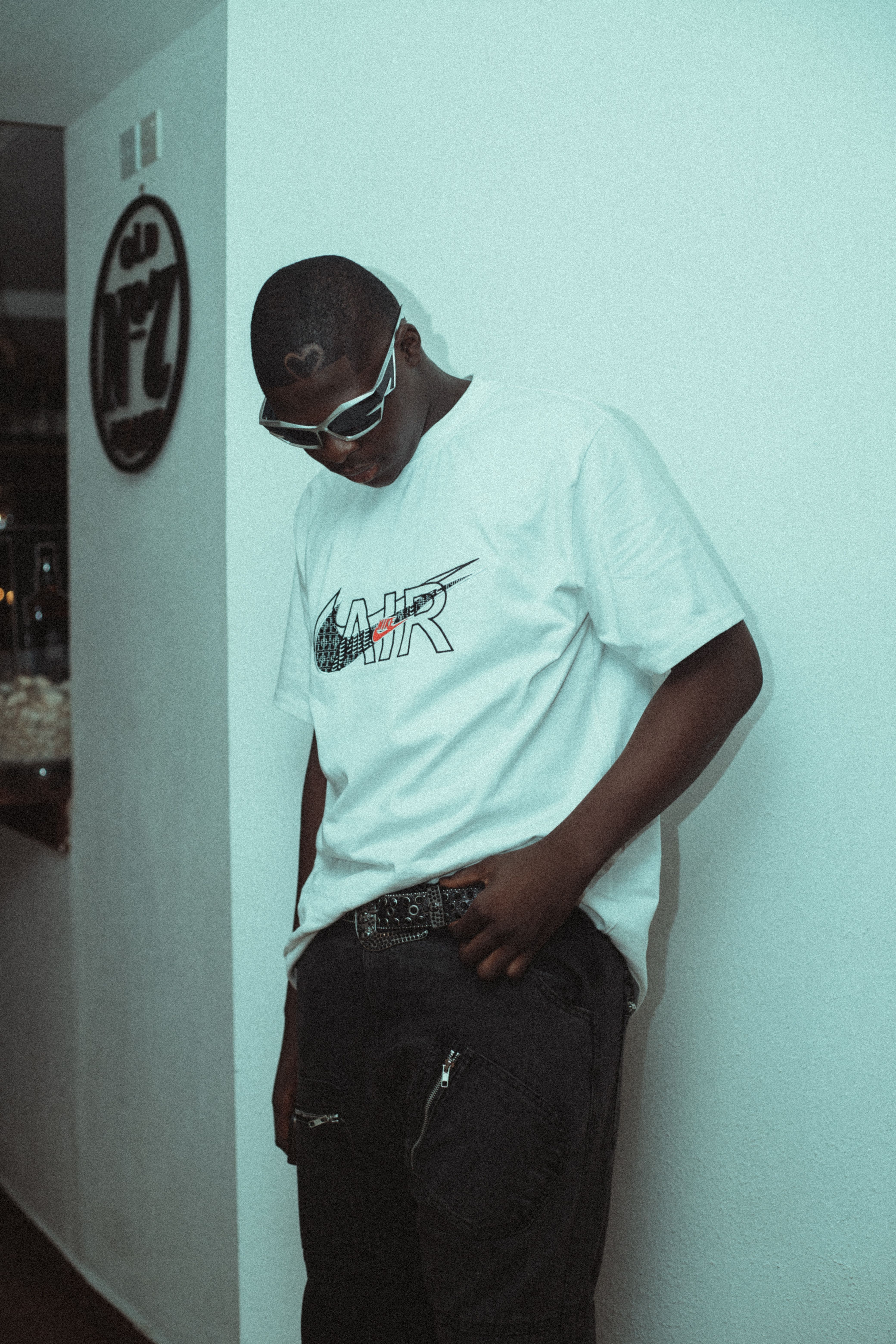Stylish person in urban apparel and sunglasses leaning against a wall indoors.
