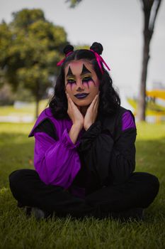 Woman in vibrant clown makeup with purple accents sitting outdoors on grass.