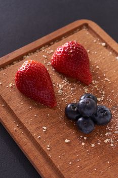 Two strawberries and blueberries arranged on a wooden cutting board with crumb sprinkles.