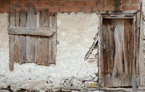 Old wooden gate and window on a weathered stone wall with exposed bricks, showcasing rustic and vintage architecture.