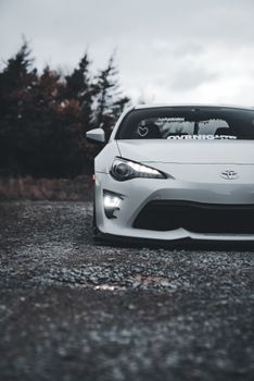 A modern white sports car parked on a gravel path lined with trees, showcasing a moody, rural setting.