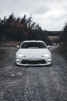 A sleek white sports car parked on a winding gravel road with a forest backdrop.