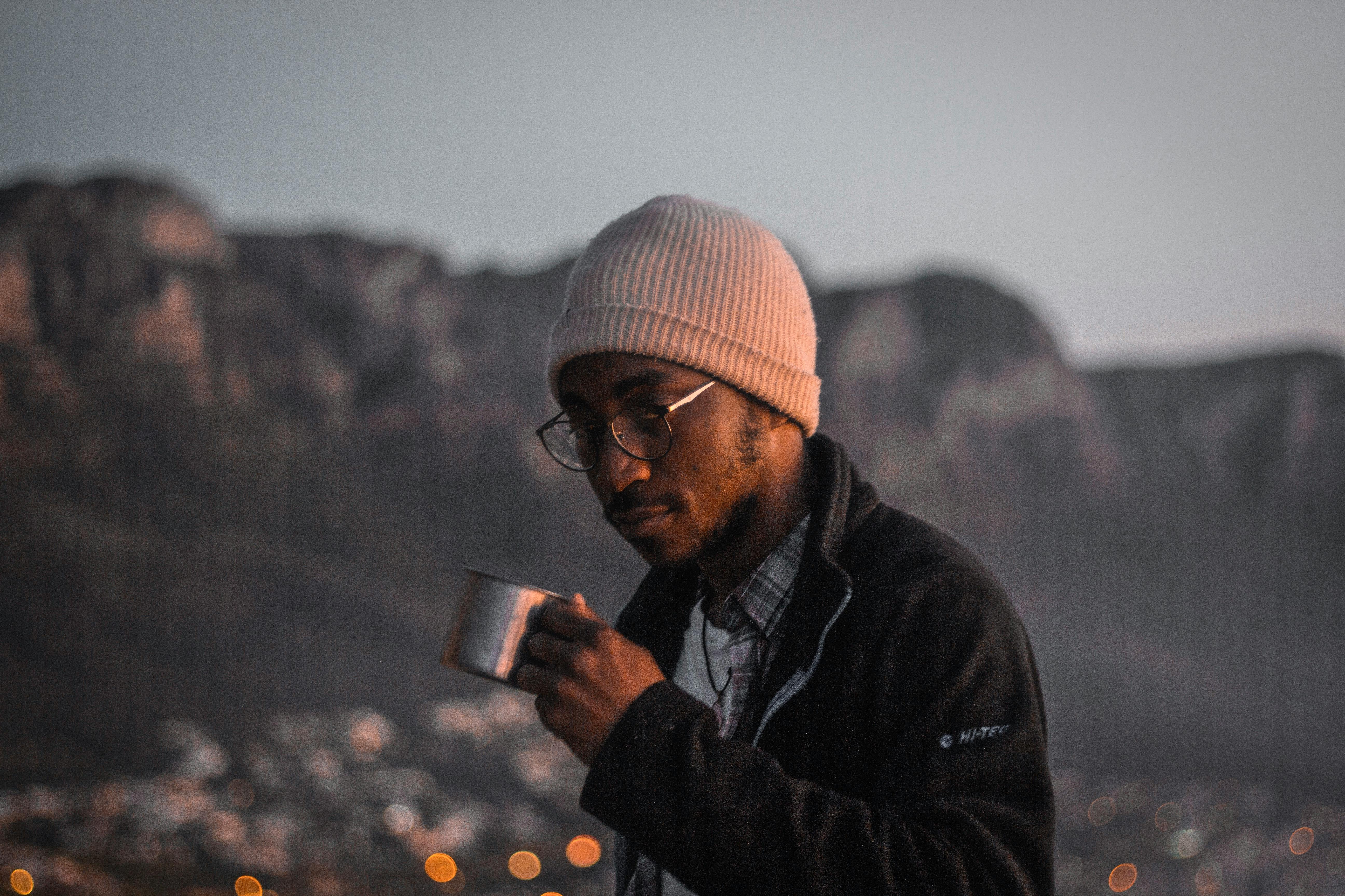 A man in a knit cap drinks from a metal cup at twilight amidst a mountain backdrop.