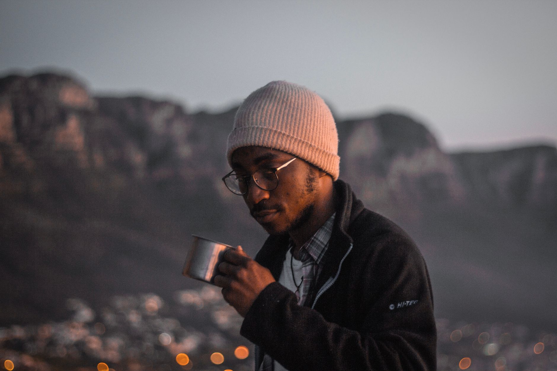 A man in a knit cap drinks from a metal cup at twilight amidst a mountain backdrop.