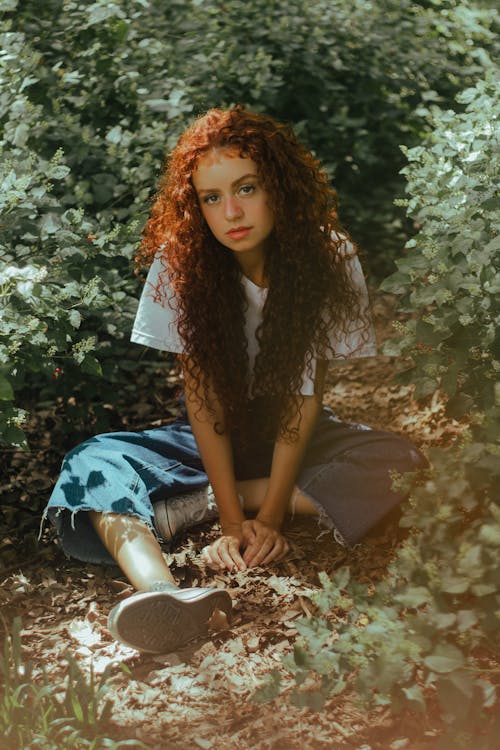 Женщина с вьющимися волосами сидит на земле возле зеленых листьев