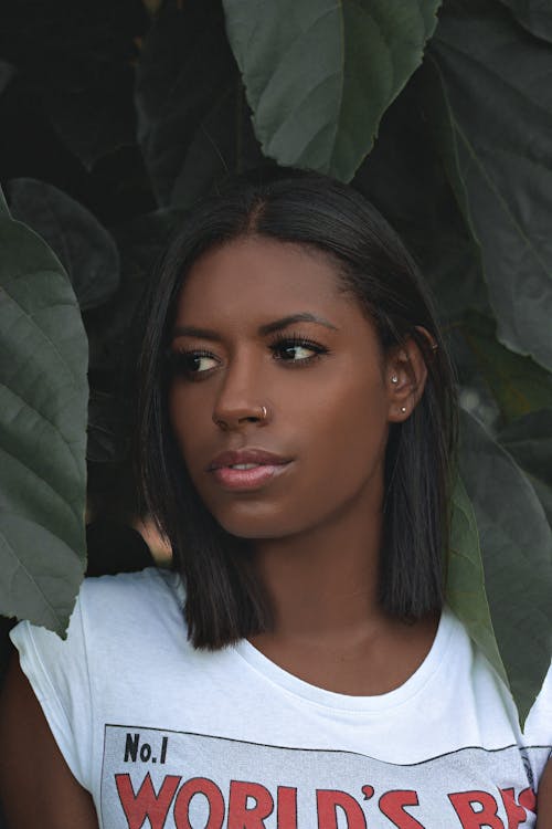 Foto De Uma Mulher Em Uma Camiseta Branca Cercada Por Plantas De Folhas Verdes Posando Enquanto Olha Para Longe