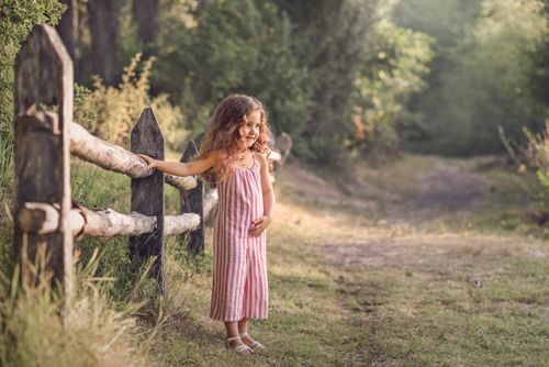 Free A smiling girl in a striped dress enjoying a sunny day by a wooden fence in nature. Stock Photo