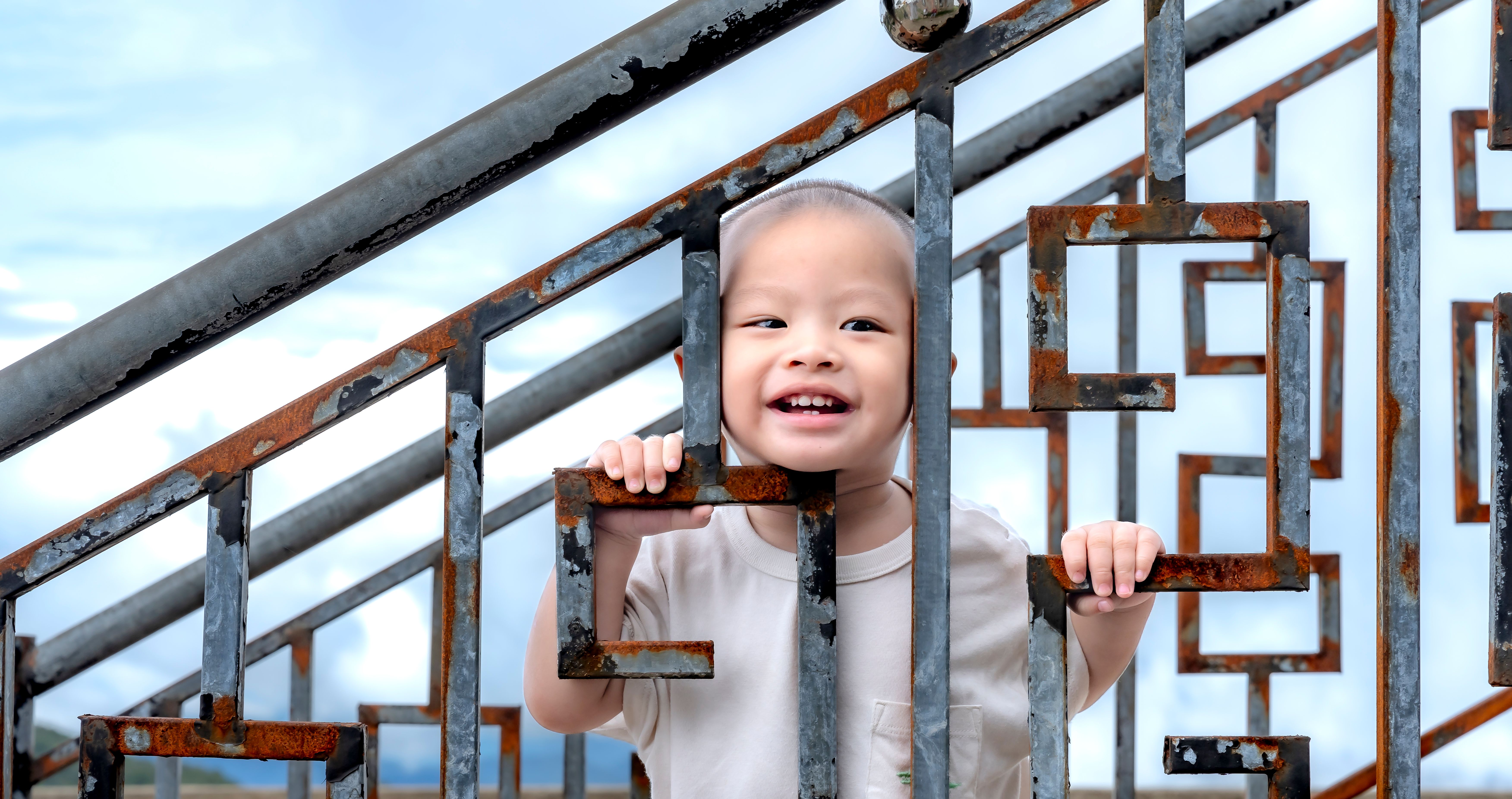 A cheerful child peeking through a rusted metal fence outdoors against a blue sky.