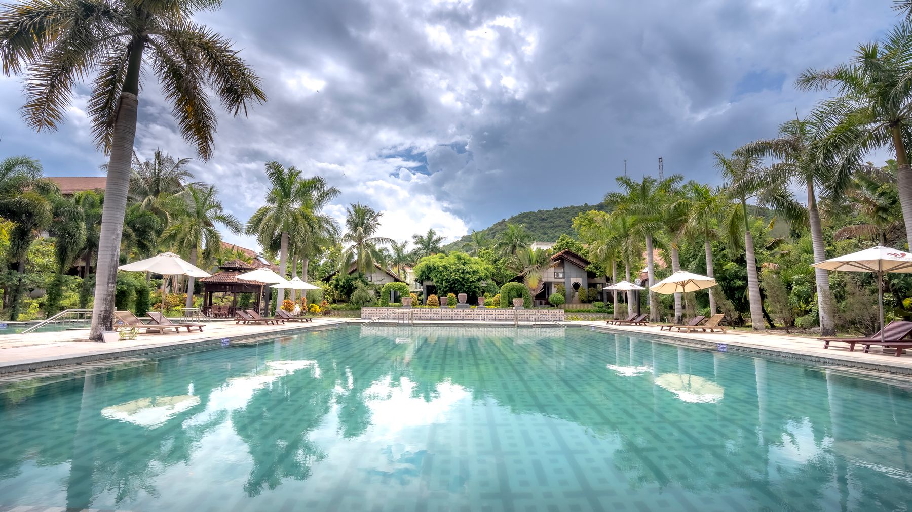 Scenic view of a tropical resort pool surrounded by palm trees under cloudy skies.