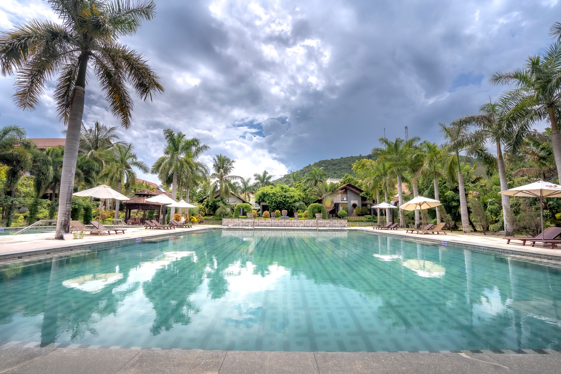 Scenic view of a tropical resort pool surrounded by palm trees under cloudy skies.