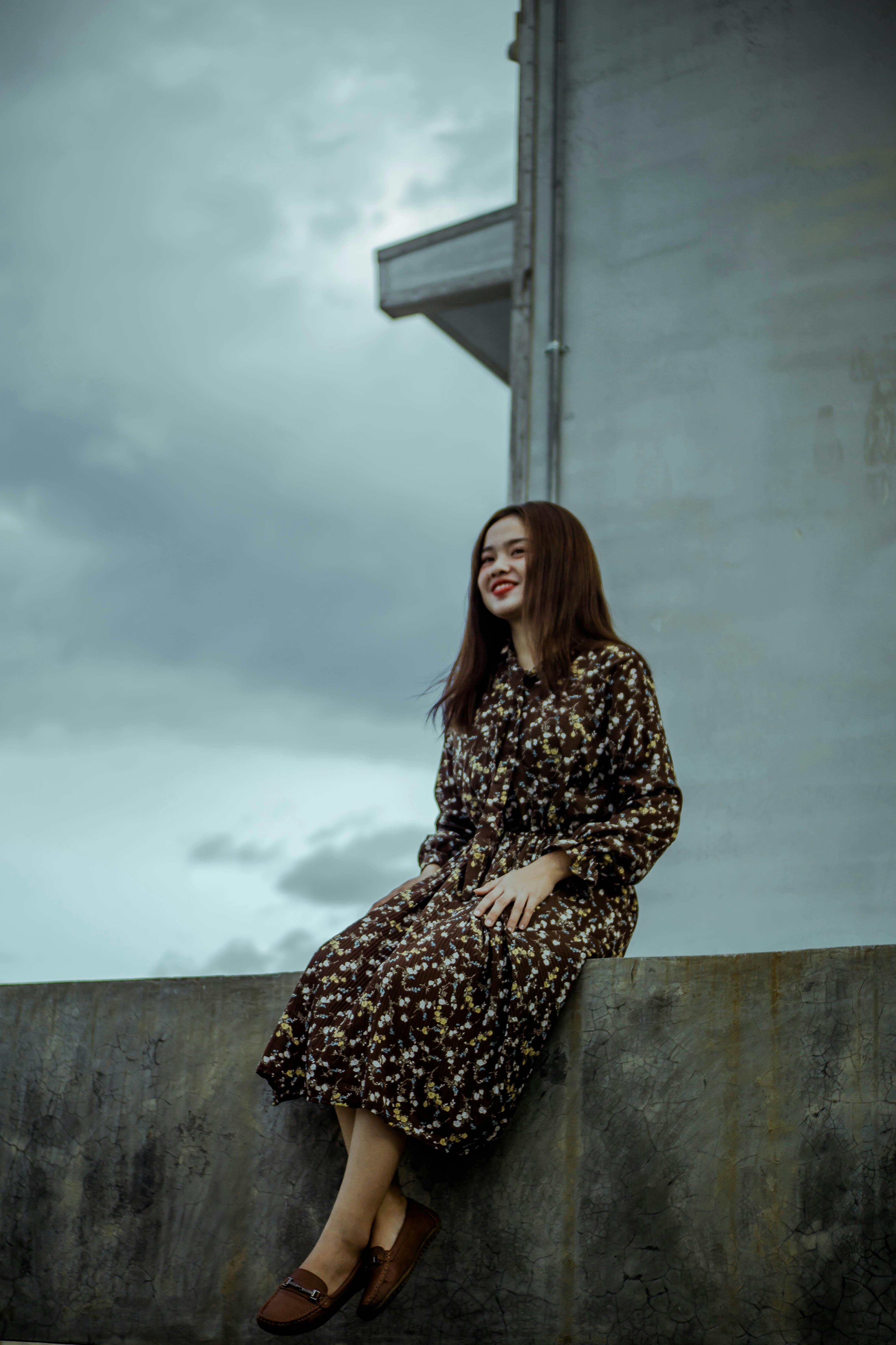 天空多云，一名身穿深色花卉连衣裙的女子坐在混凝土墙上微笑着