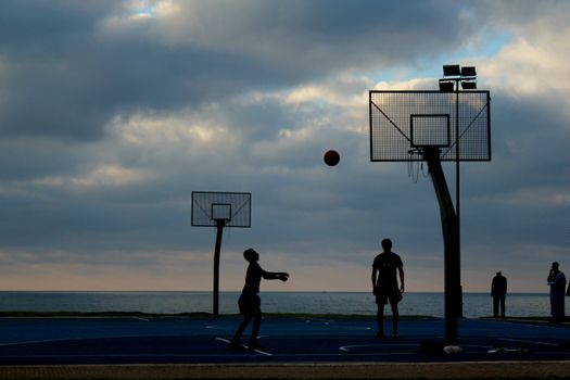 Basketball players silhouetted at a seaside court during sunset.