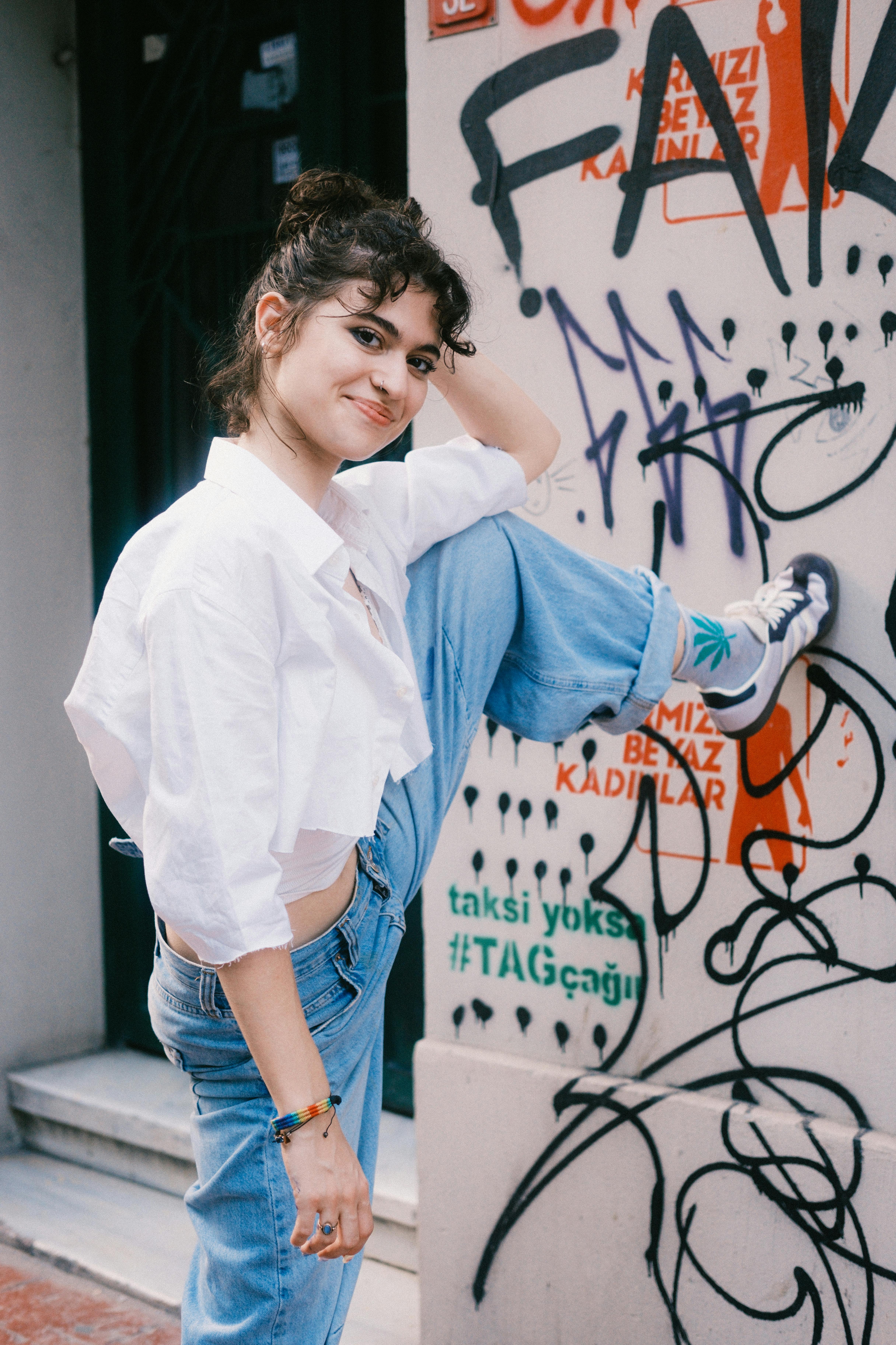 Young woman in casual attire poses with leg raised next to vibrant graffiti wall. Urban and playful vibe.