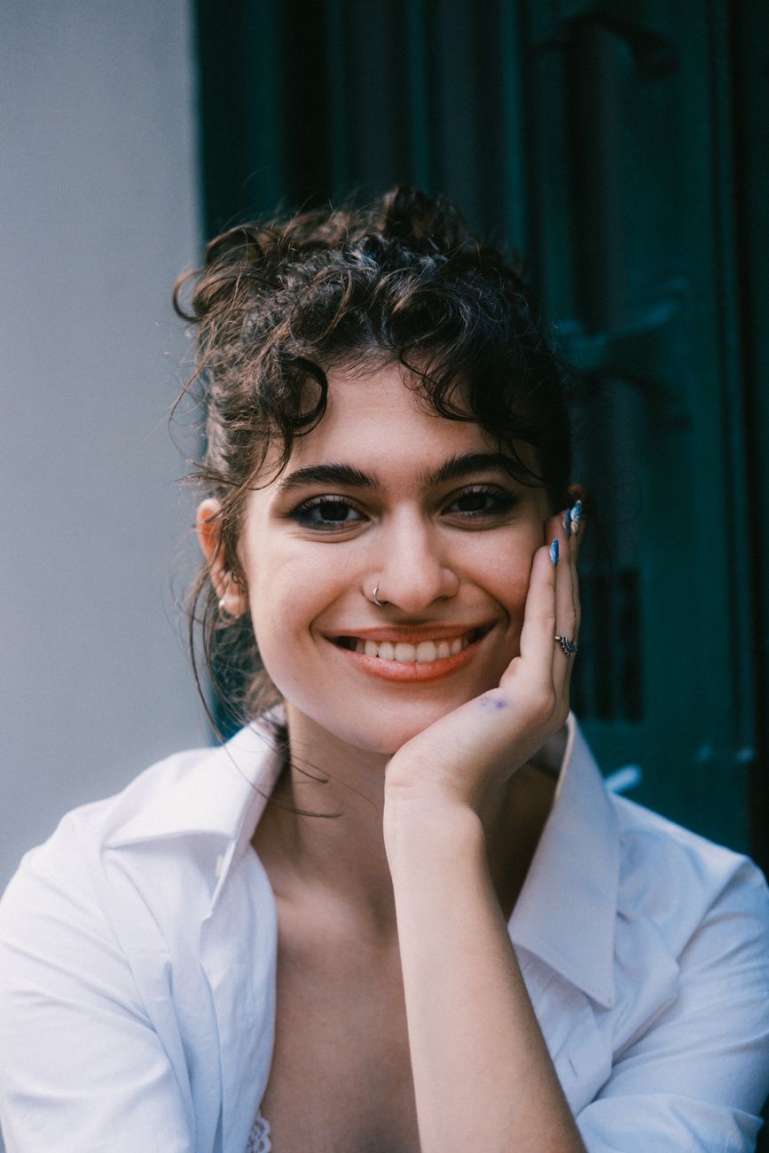 Young woman with curly hair smiling, wearing a white shirt outdoors.