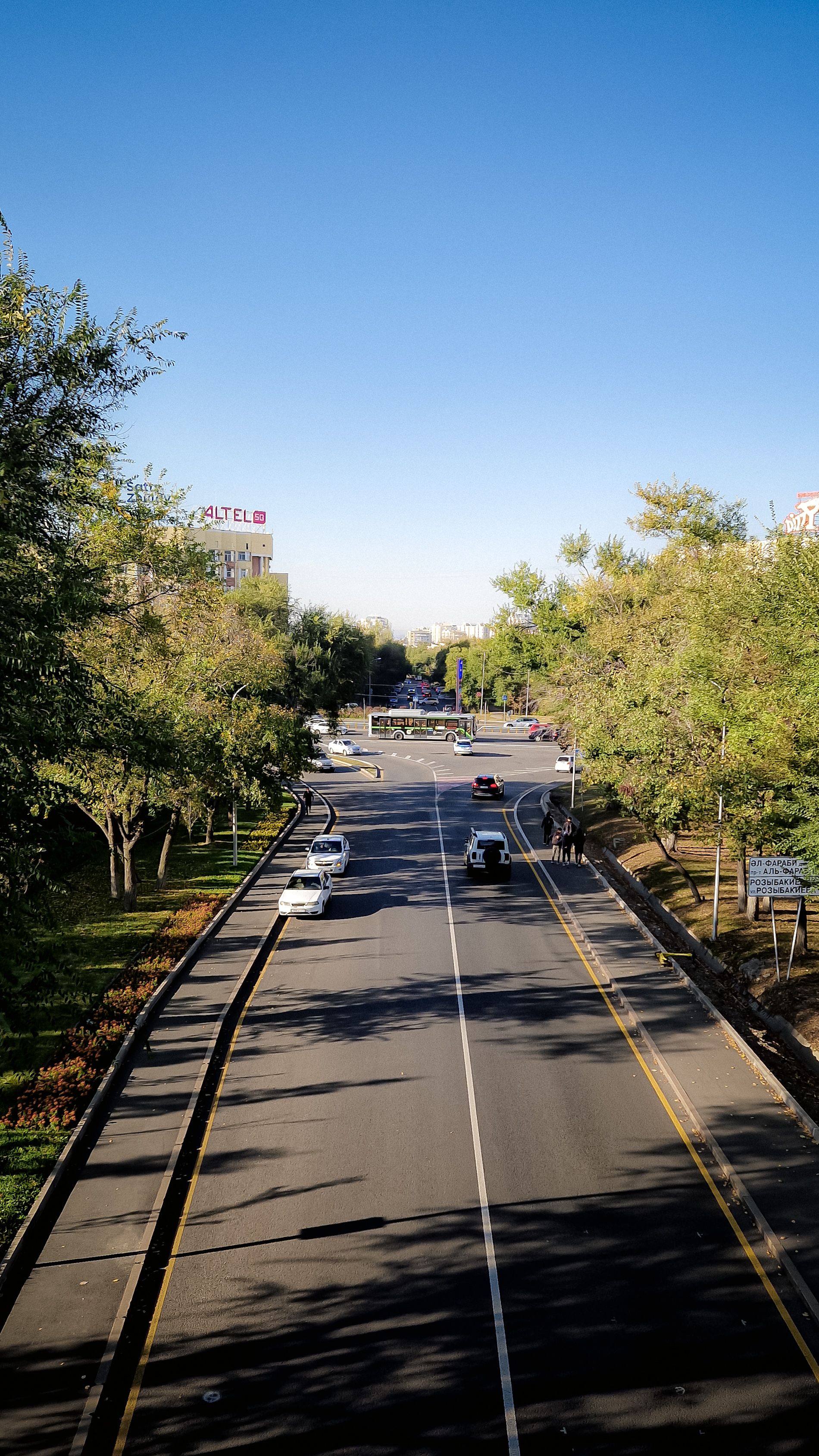 Aerial view of a city street lined with trees and moving vehicles under a clear blue sky.