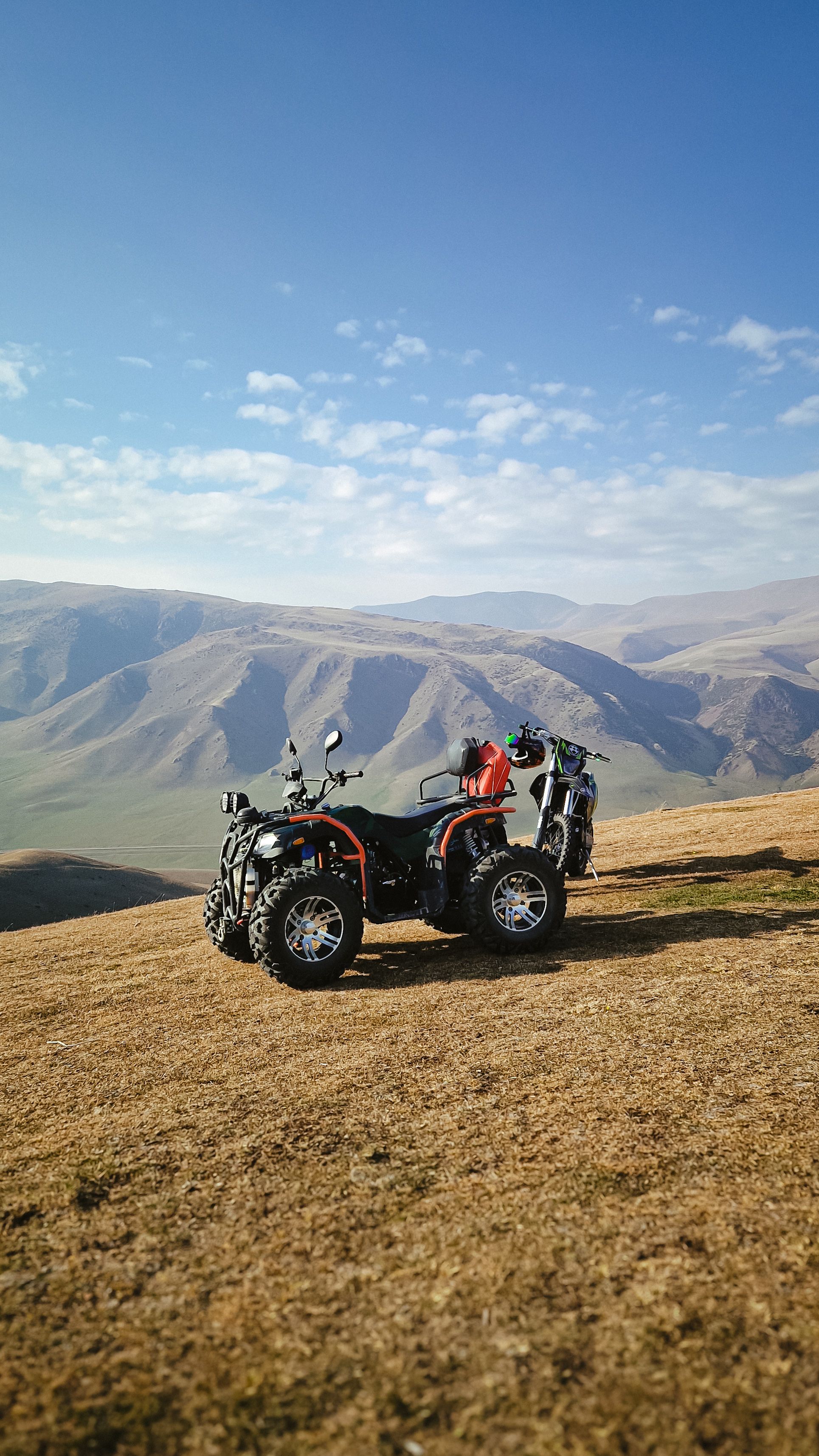 ATV parked on a grass-covered hill with a panoramic mountain view under a blue sky.