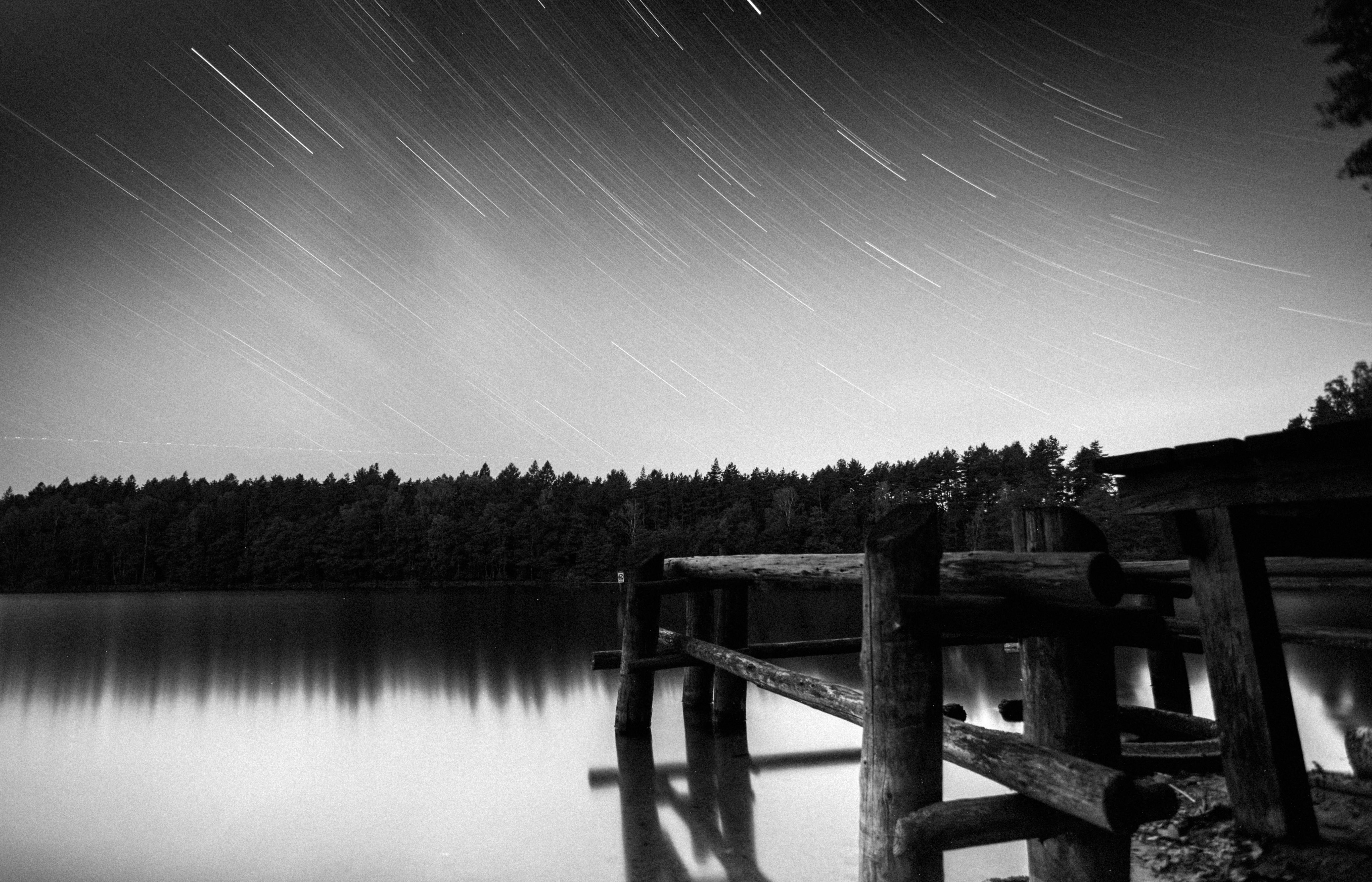 Black and white shot of a starry night over a serene lake in Poland, featuring a wooden pier and forest backdrop.