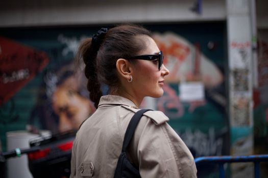 Stylish woman wearing sunglasses and a trench coat in front of urban graffiti.