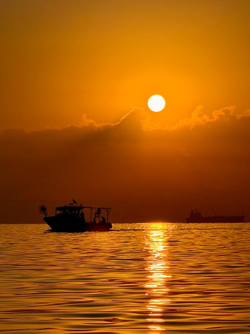 Free A silhouette of a boat at sunset, reflecting on calm ocean waters, creating a tranquil scene. Stock Photo