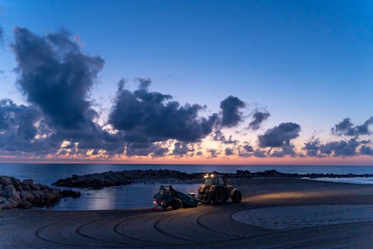 A tractor working at sunrise on a beach in Comunidad Valenciana, Spain, with dramatic clouds overhead.
