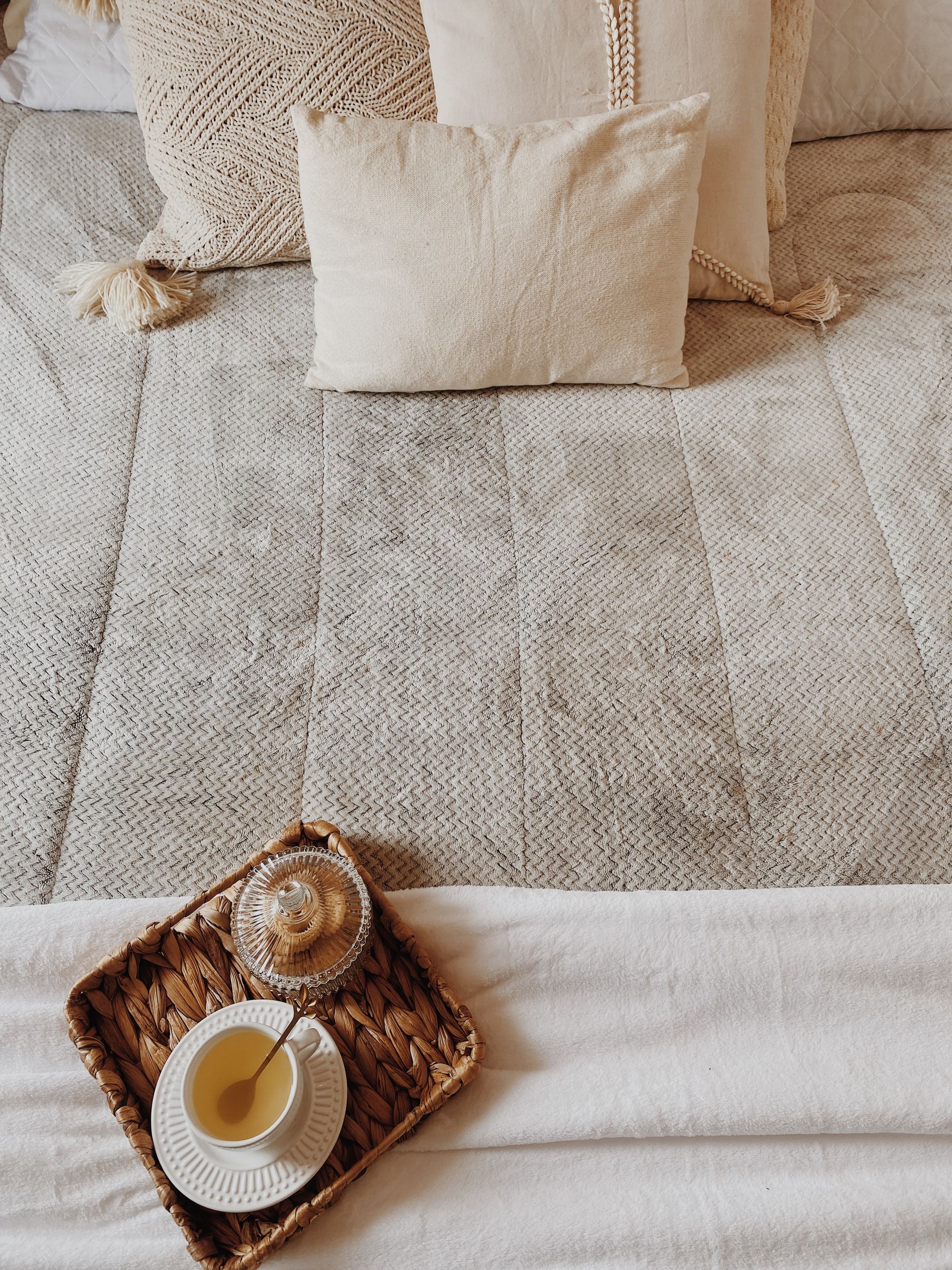 Cream and beige bedding layered with woven cushions, tassel trim and a wooden tray