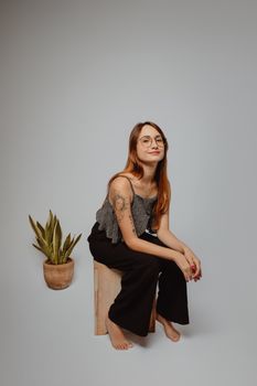 Young woman sitting indoors with a plant, showcasing modern fashion and casual style.